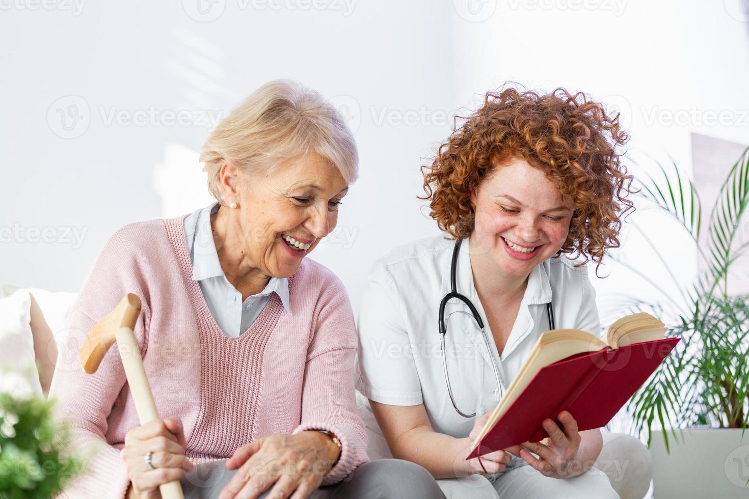 Frauenbetreuerin, die ein Buch liest, während sie mit einer glücklichen Seniorin im Pflegeheim sitzt. glückliche ältere frau, die auf einem weißen sofa sitzt und der krankenschwester zuhört, die ein buch laut vorliest foto