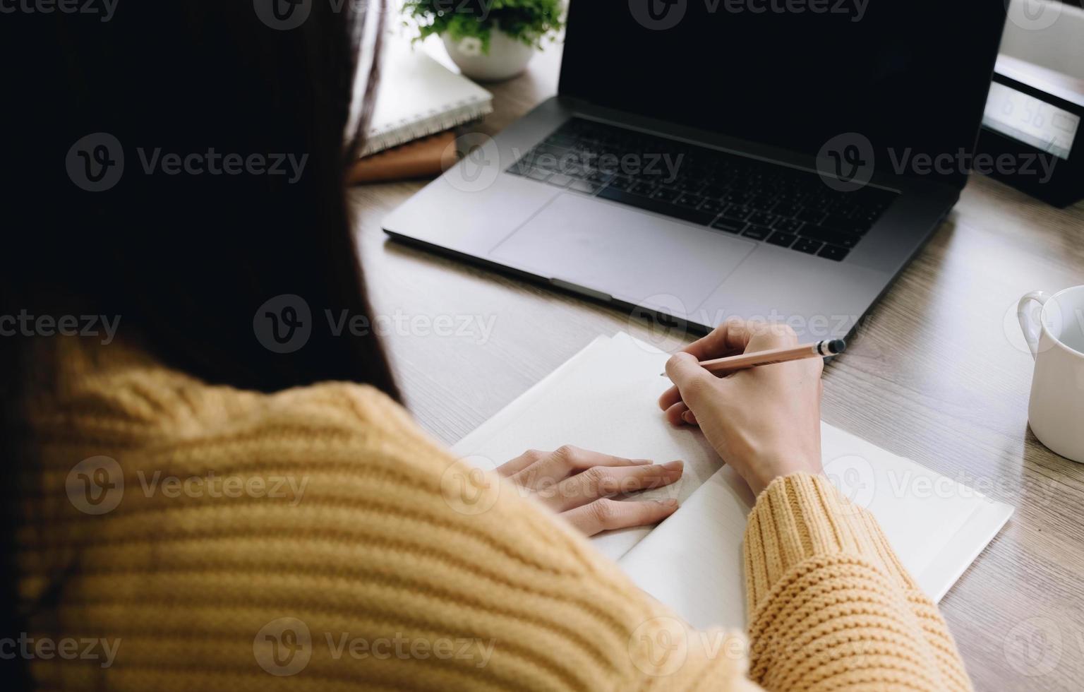 Frau schreibt Notizen, während sie vor ihrem Computer-Laptop am hölzernen Arbeitstisch über dem Wohnzimmer sitzt foto