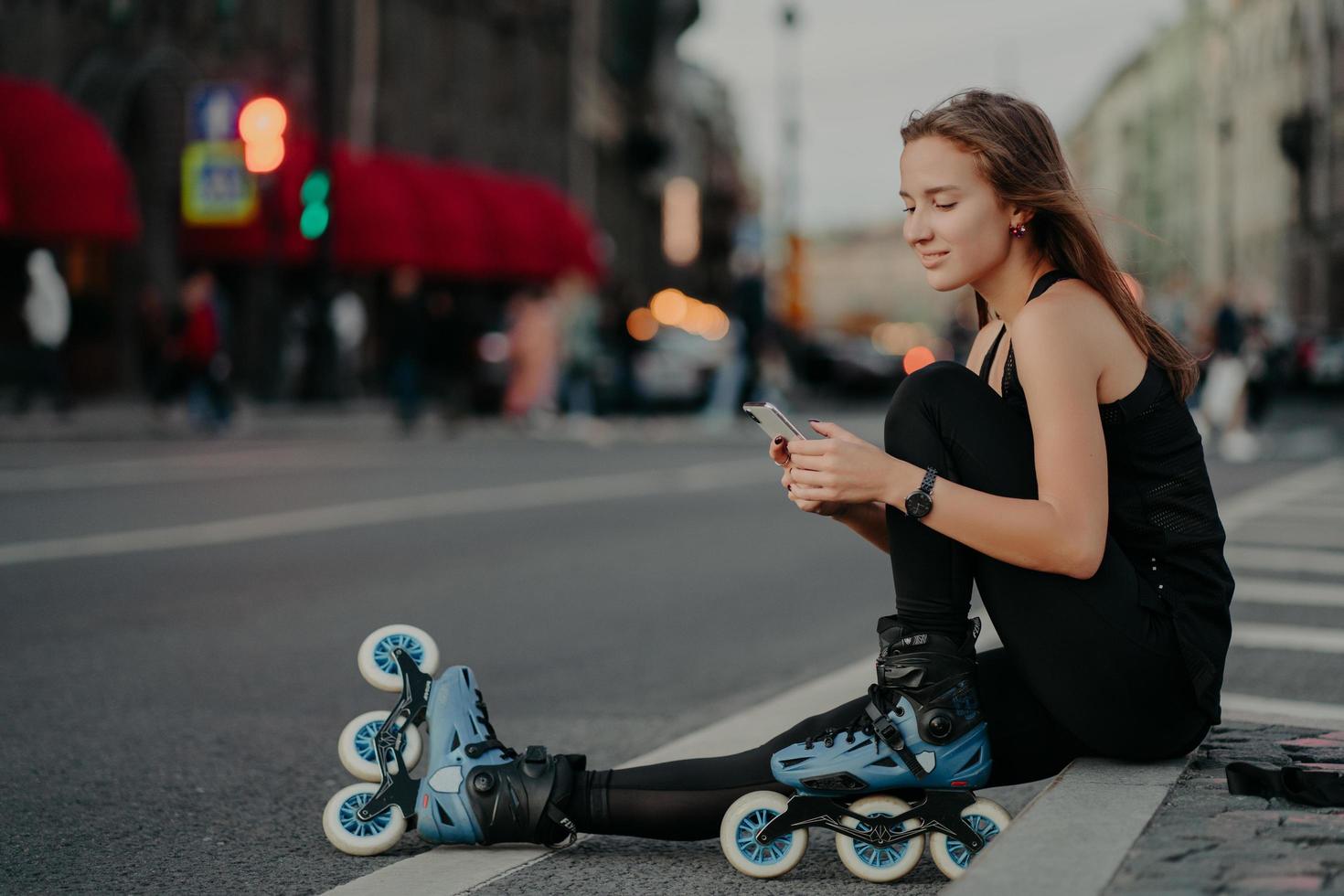 Außenaufnahme einer sportlich aktiven jungen Frau in aktiver Kleidung trägt Inlineskates und überprüft Nachrichteninhalte auf Smartphone-Posen vor verschwommenem Stadthintergrund auf Asphalt führt zu einem gesunden Lebensstil foto