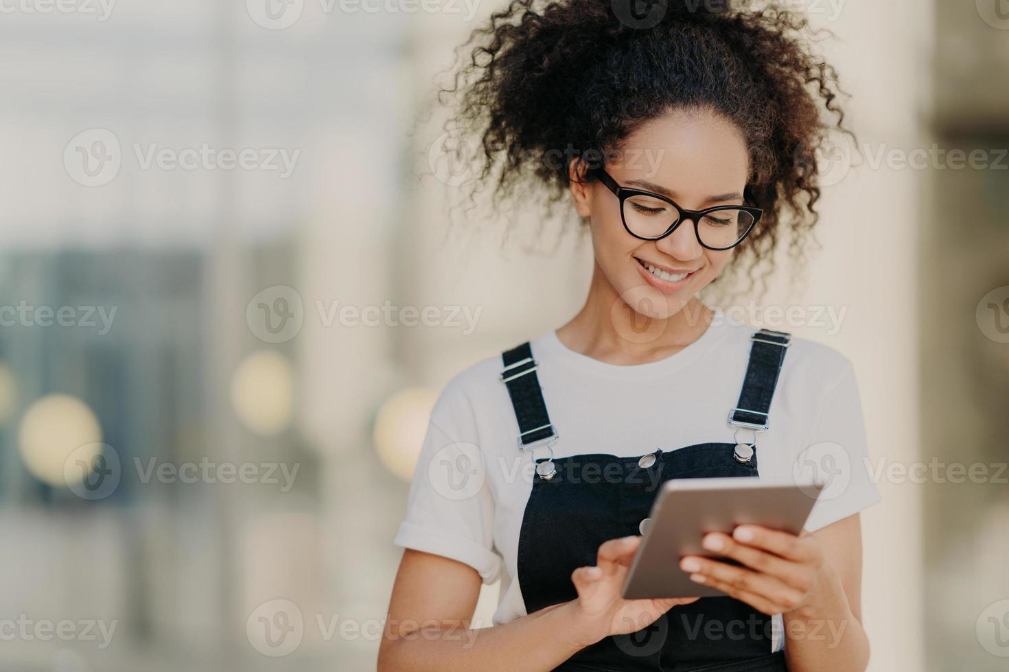 Foto eines attraktiven tausendjährigen Mädchens mit krausem Haar, hält ein digitales Tablet in der Hand, surft in sozialen Netzwerken, trägt eine optische Brille, ein lässiges T-Shirt und eine Sarafan, ist mit dem drahtlosen Internet verbunden und gibt eine Nachricht ein