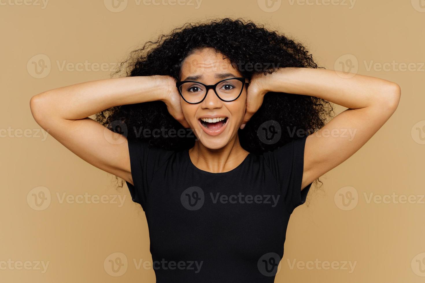 junge frau mit lockigem afro-haar bedeckt ohren, schreit, wenn sie verlangt, leise zu sein, kann geräusche nicht ertragen, hält den mund offen, trägt eine brille und ein lässiges schwarzes t-shirt, isoliert über braunem studiohintergrund. foto