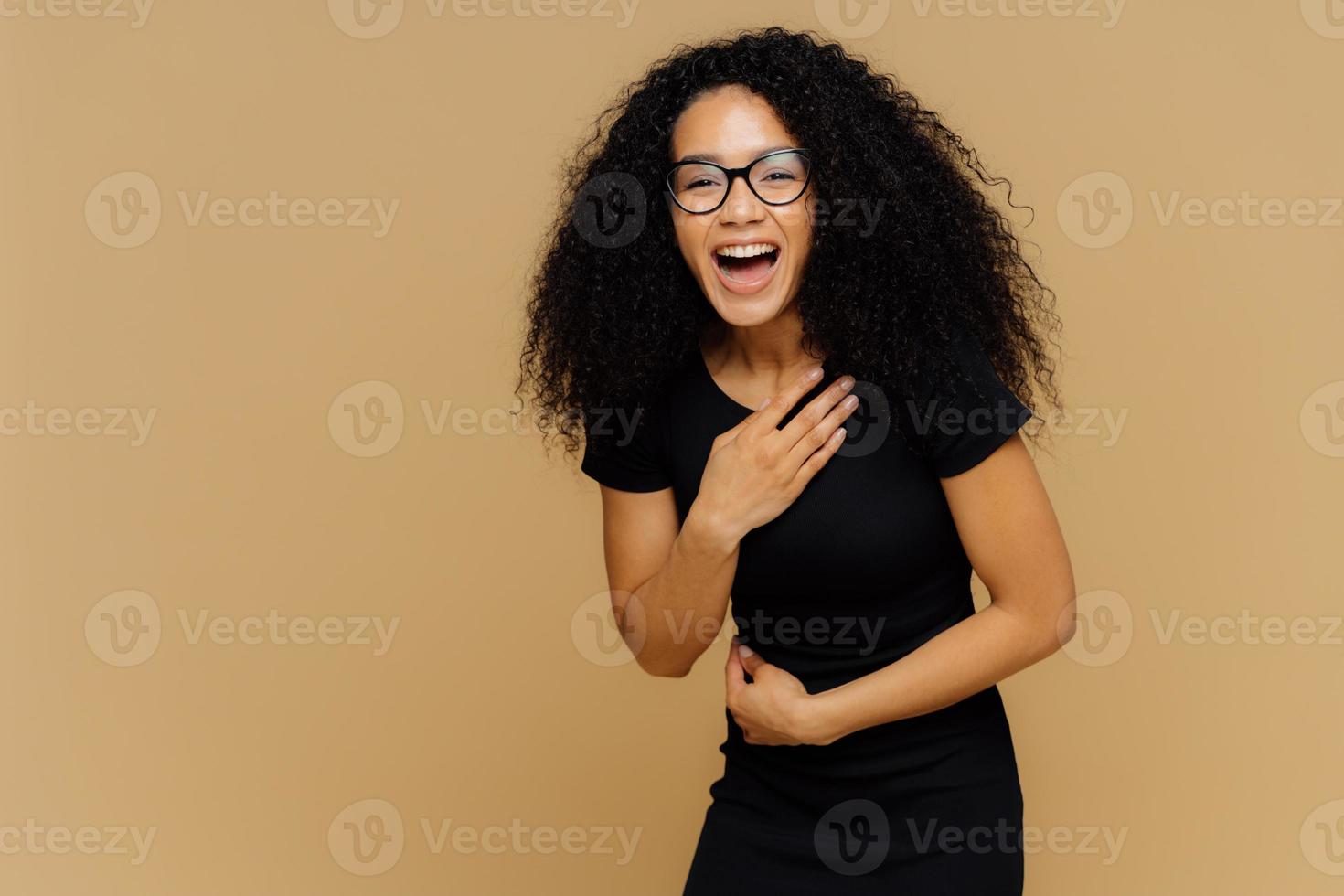 schönes dunkelhäutiges model kann nicht aufhören zu lachen, hört comicgeschichte, hält hand auf bauch, lacht laut, in schwarze kleidung gekleidet, steht an brauner studiowand mit leerem platz für informationen foto