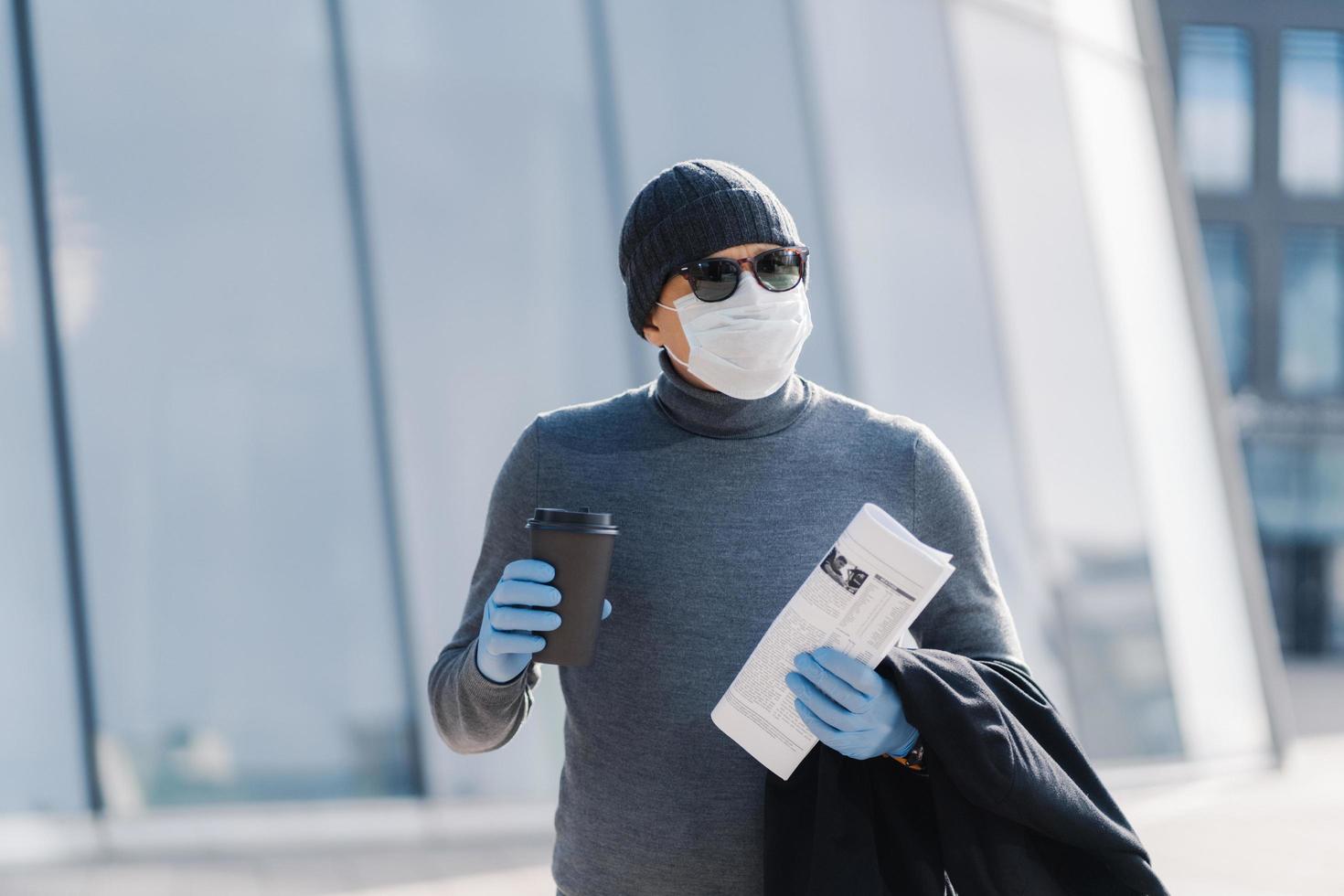 Bild des jungen Mannes trägt sterile Maske und medizinische Gummihandschuhe, schaut zur Seite, geht während der Ausbreitung von Infektionskrankheiten durch die Stadt, trinkt Kaffee, gefaltete Zeitung. Coronavirus-Präventionskonzept foto