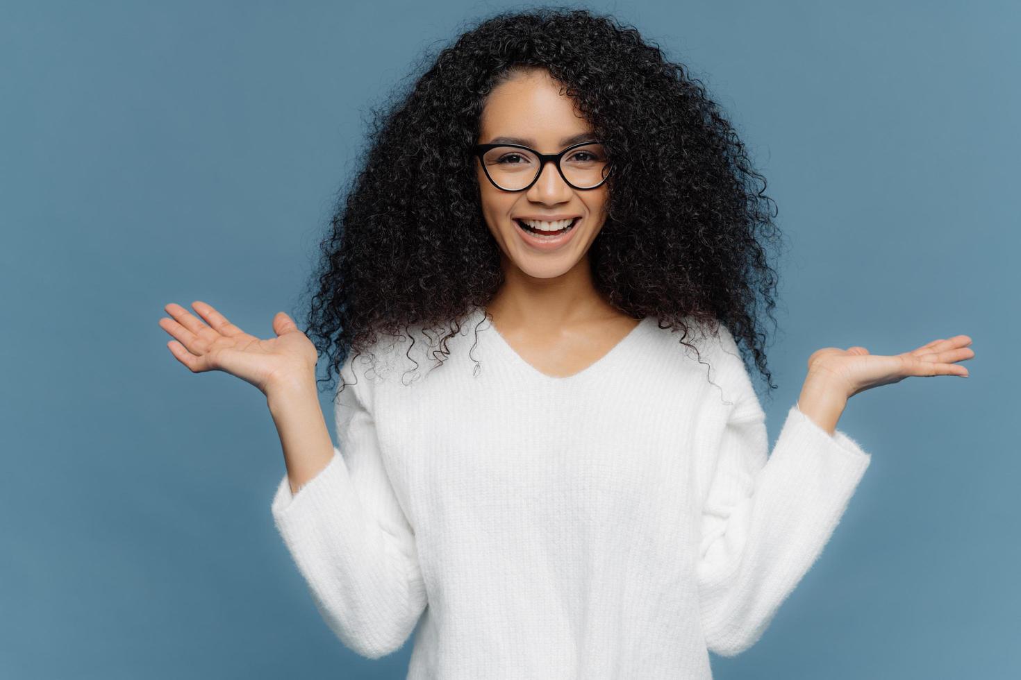 positive freundliche junge frau mit afro-aussehen, breitet handflächen aus, hält unsichtbares objekt, hat charmantes lächeln, gesunde haut, trägt brille und weißen pullover, isoliert auf blauem hintergrund. foto