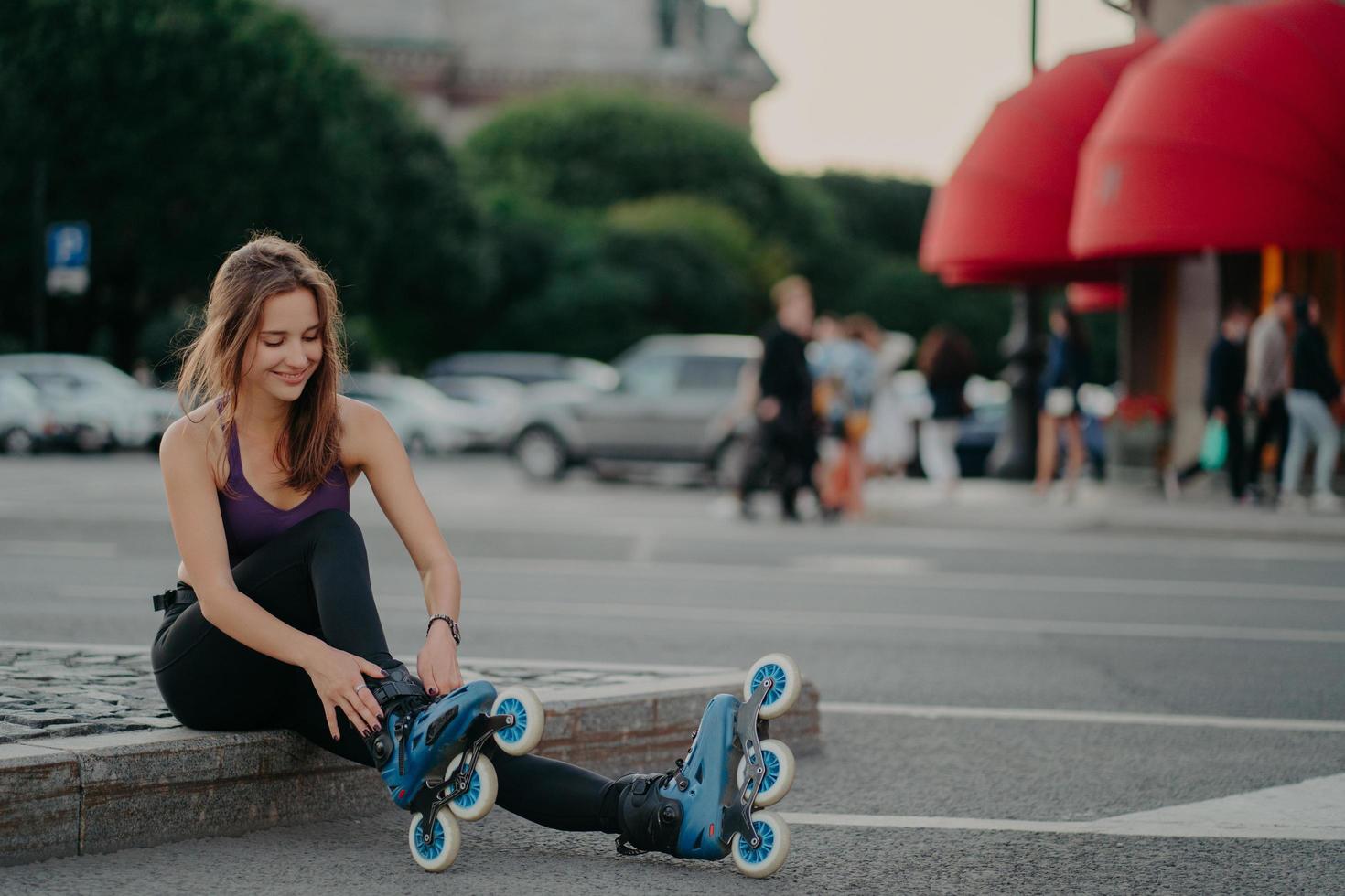sportlicher Lifestyle und Hobby. Zufriedene dunkelhaarige Europäerin zieht Inline-Skates an und geht Inline-Skating-Posen vor verschwommenem Stadthintergrund, hält sich fit und verbringt ihre Freizeit aktiv. Außenaufnahme foto