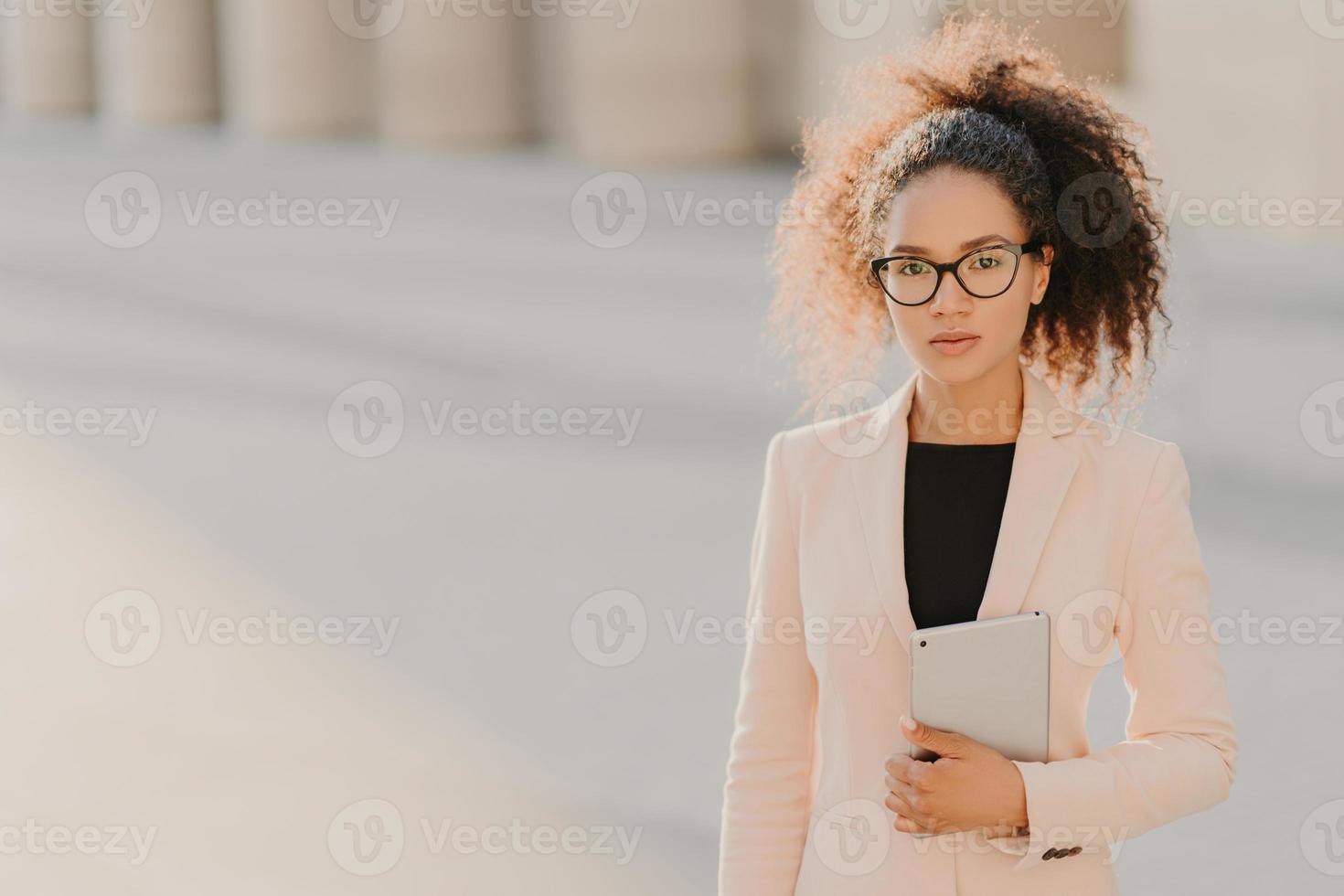 Foto einer eleganten afroamerikanischen Geschäftsfrau verwendet einen Tablet-Computer im Freien, trägt eine formelle weiße Jacke, eine transparente Brille mit schwarzem Rahmen, lockiges Haar zu einem Pferdeschwanz gekämmt, eine Leerstelle auf der linken Seite