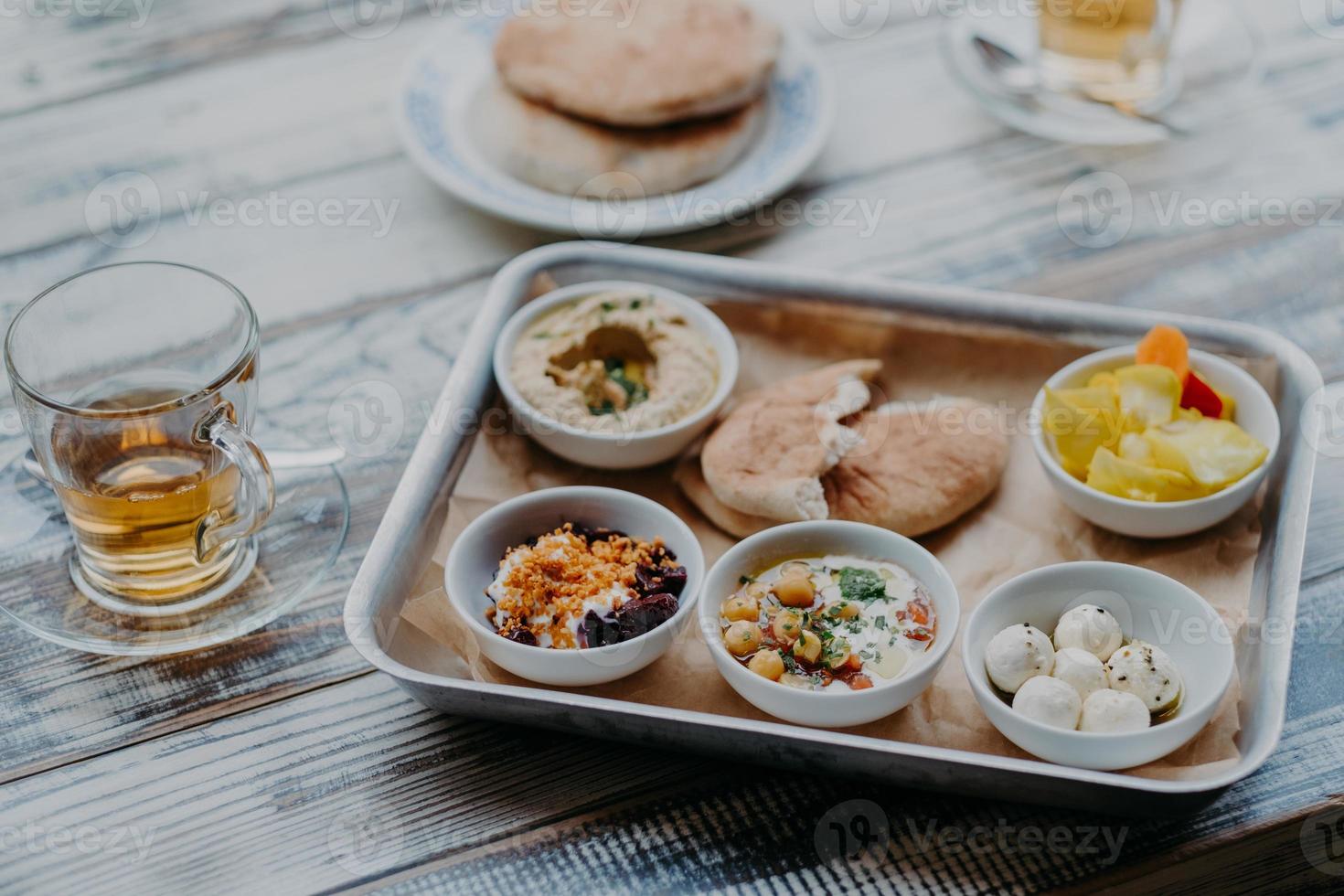 Overhead-Bild des traditionellen israelischen Essens auf dem Tablett. Hummus, heimischer Ziegenkäse, Tomatenkern, Rote Beete mit Gewürzen, Fladenbrot, Getränk im Glas foto