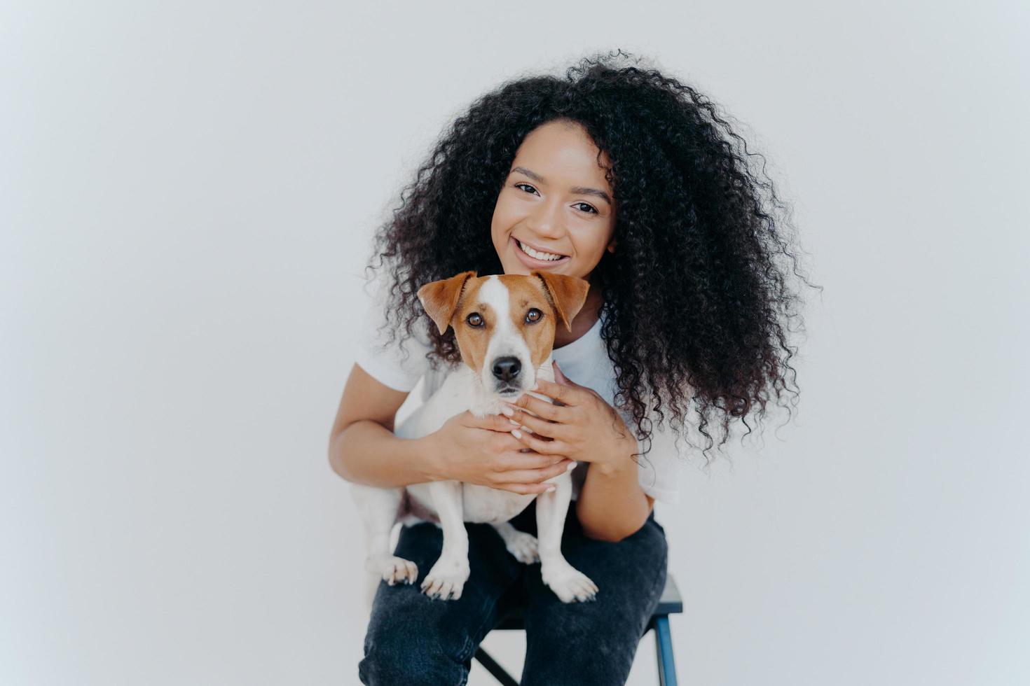 lächelnde afrikanisch-amerikanische frau posiert mit haustier, schaut freudig in die kamera, kuschelt hund, sitzt auf stuhl, isoliert über weißem hintergrund. zwei Familienmitglieder. gemütlich entspannen. für immer zusammen foto