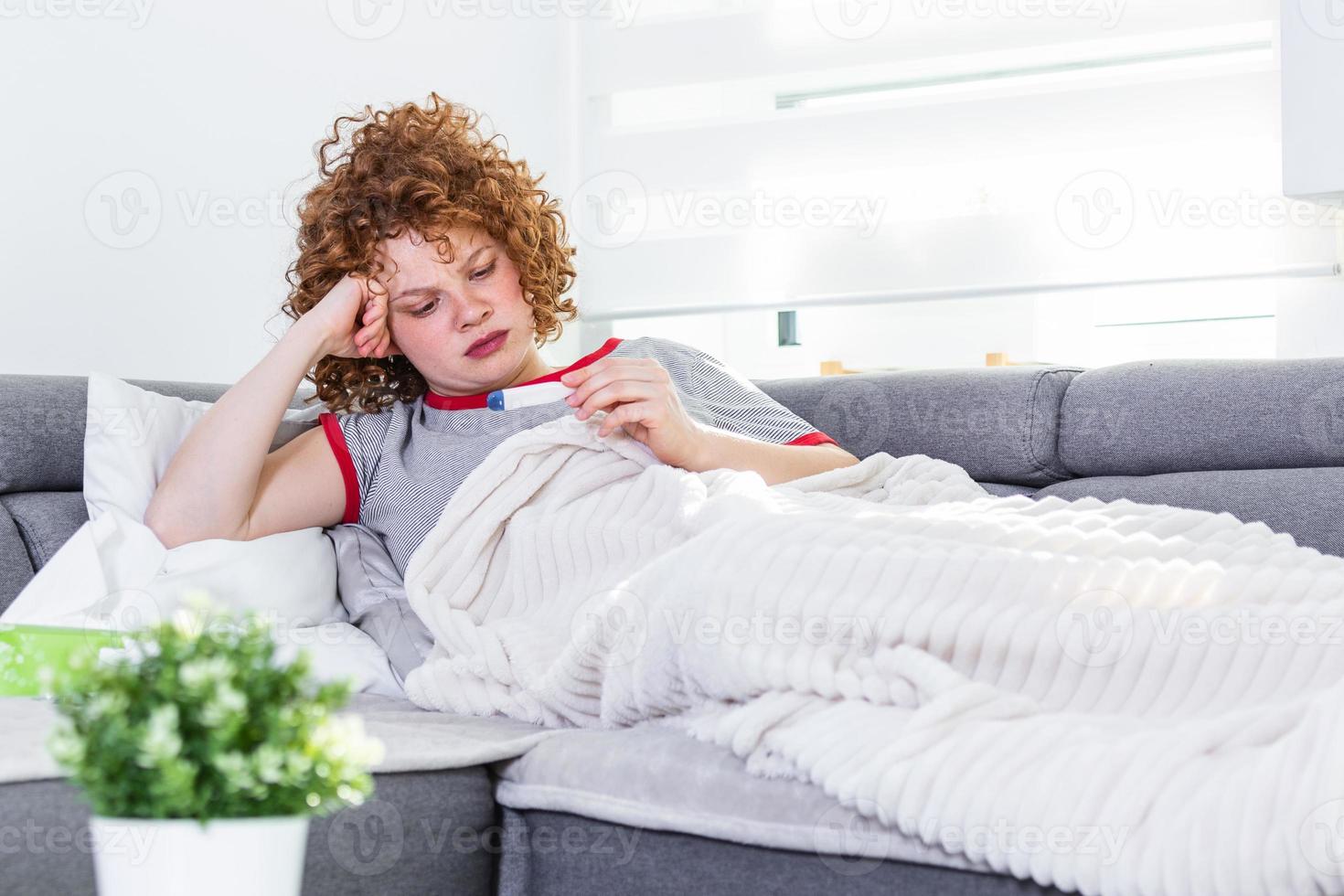 krankheit, saisonales virusproblemkonzept. Frau, die krank ist und eine Grippe hat, die auf dem Sofa liegt und die Temperatur auf dem Thermometer ansieht. kranke frau, die mit hohem fieber im bett liegt. Erkältung und Migräne. foto