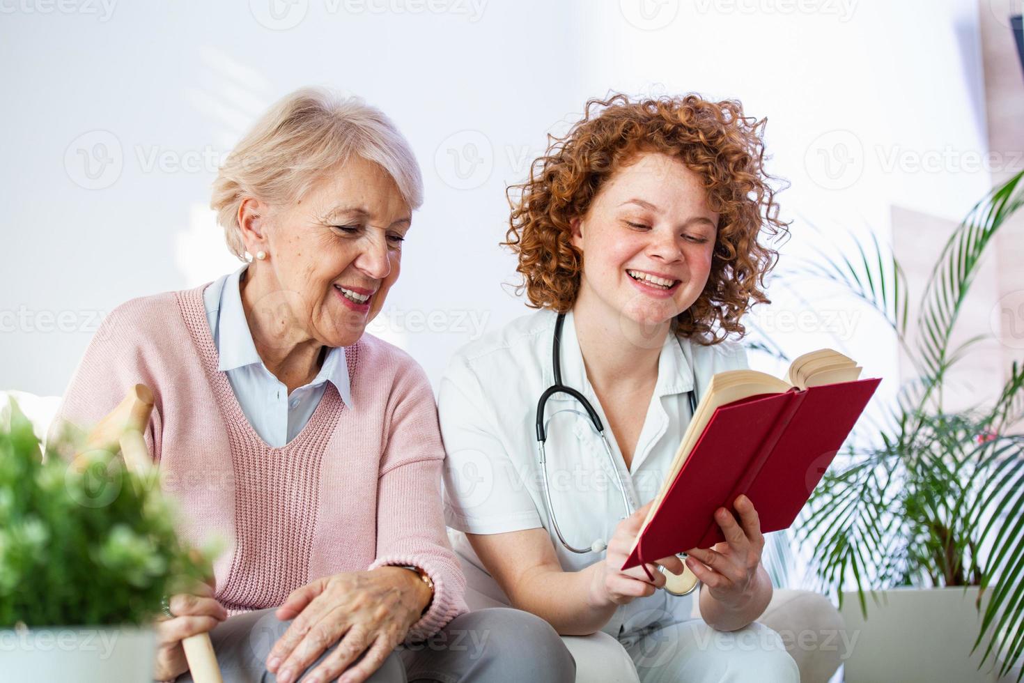 Frauenbetreuerin, die ein Buch liest, während sie mit einer glücklichen Seniorin im Pflegeheim sitzt. glückliche ältere frau, die auf einem weißen sofa sitzt und der krankenschwester zuhört, die ein buch laut vorliest foto