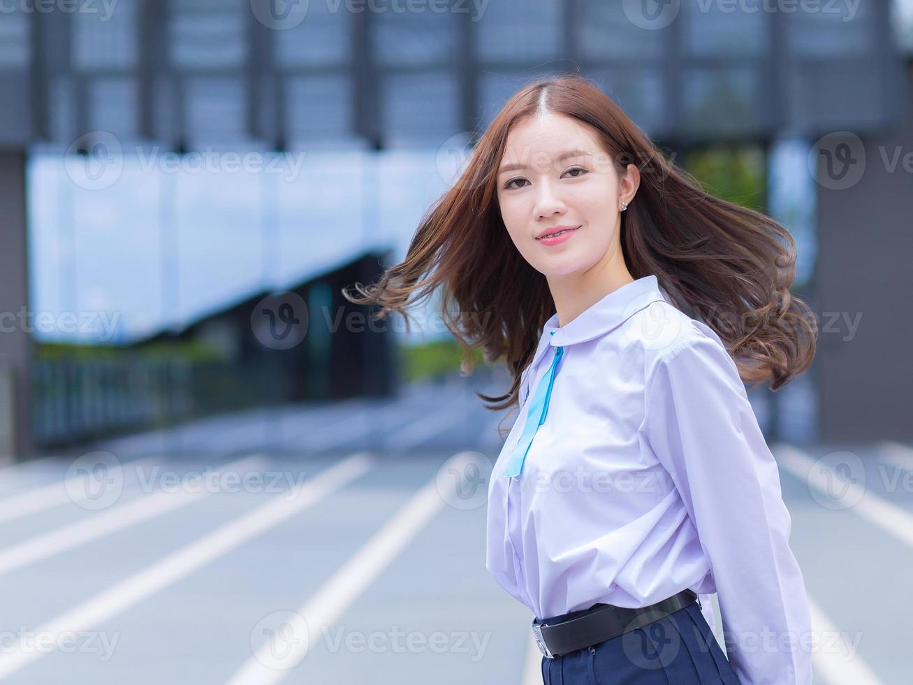 Das hübsche asiatische Highschool-Mädchen in Schuluniform mit Hosenträgern dreht sich um, um an einem hellen Tag mit dem Gebäude im Hintergrund selbstbewusst auf die Kamera zu schauen. foto