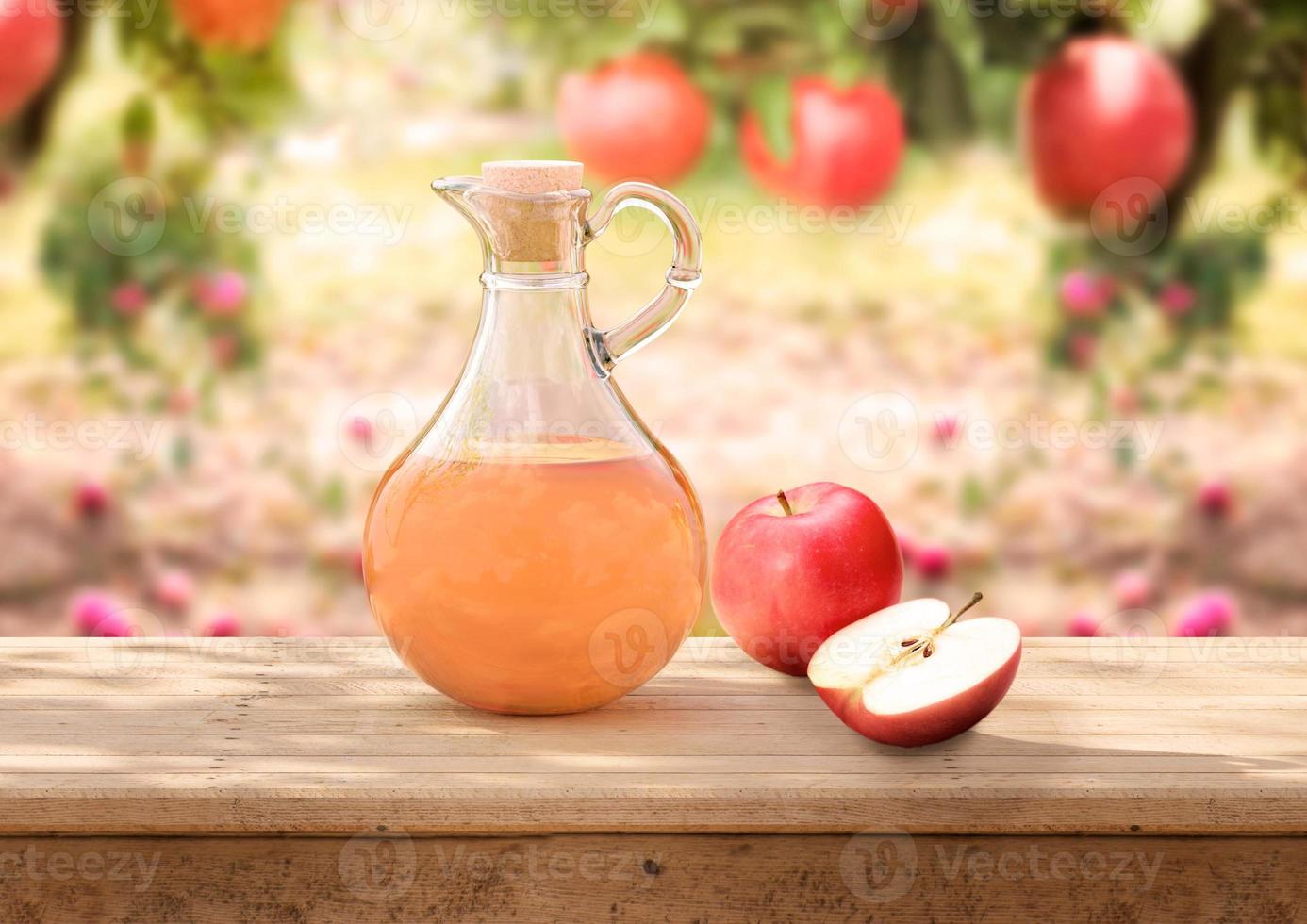 Bio-Apfelessig im Glaskrug auf rustikalem Holztisch Apfelplantage im Hintergrund foto