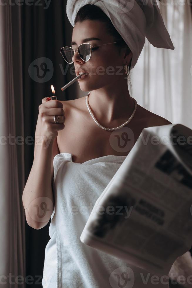Schöne junge Frau in einem Handtuch raucht eine Zigarette und liest Zeitung foto
