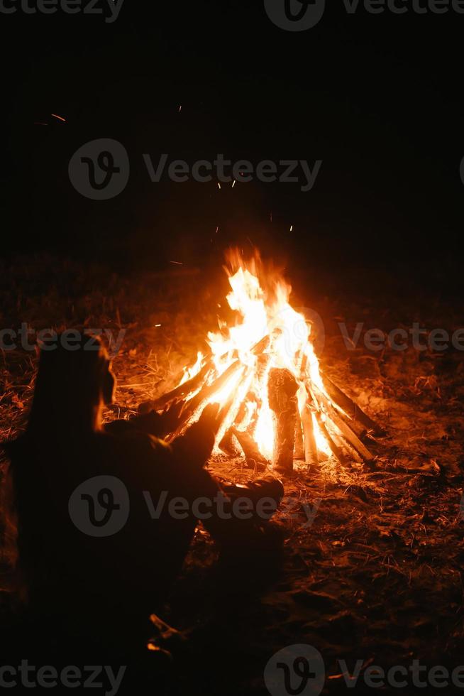 Frau, die am Lagerfeuer im Nachtwald sitzt und sich erwärmt. foto