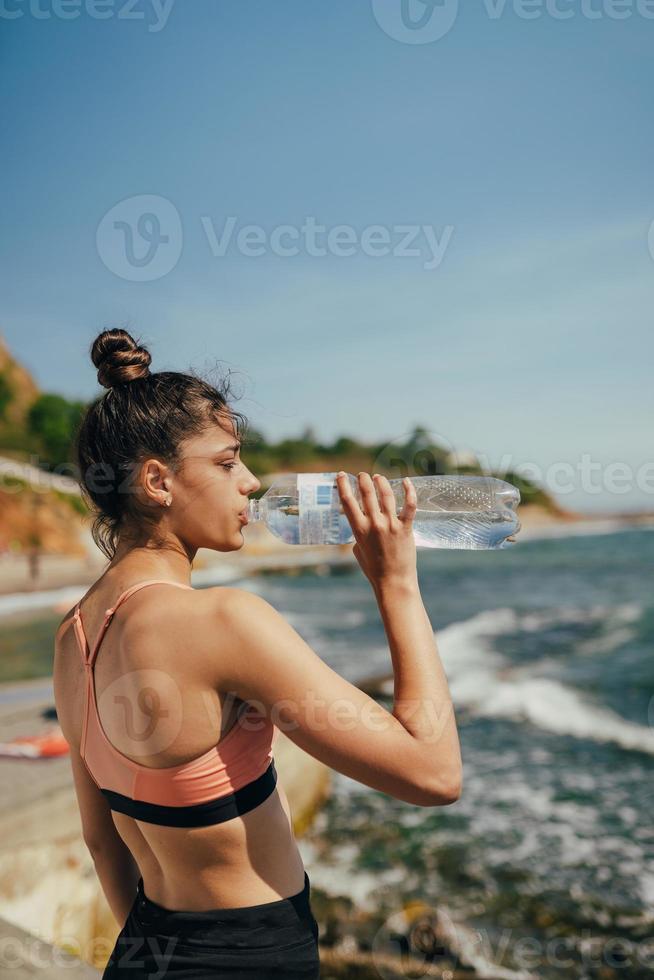 Frau, die nach dem Training am Strand frisches Wasser aus der Flasche trinkt foto