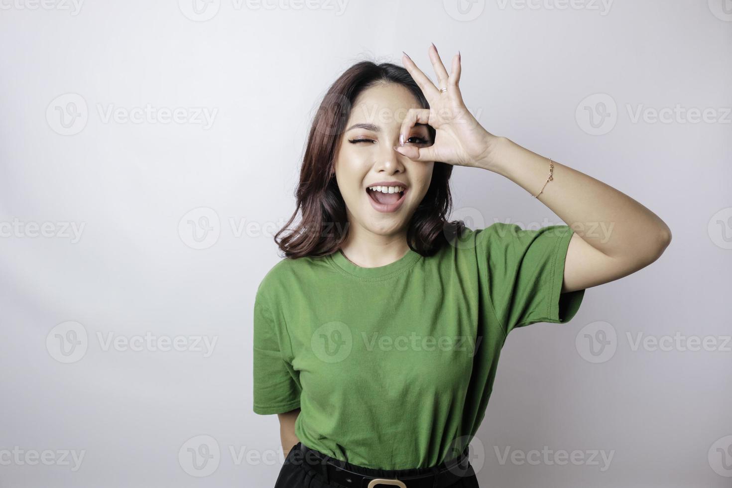 Aufgeregte asiatische Frau, die ein grünes T-Shirt trägt und eine ok-Handgeste gibt, die durch einen weißen Hintergrund isoliert wird foto