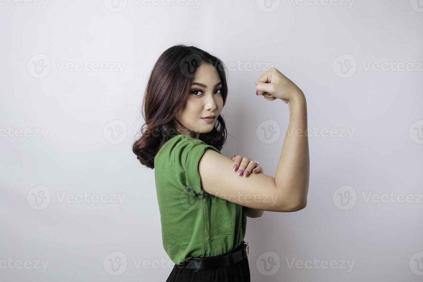 aufgeregte asiatische frau, die ein grünes t-shirt trägt, das eine starke geste zeigt, indem sie ihre arme und muskeln stolz lächelt foto