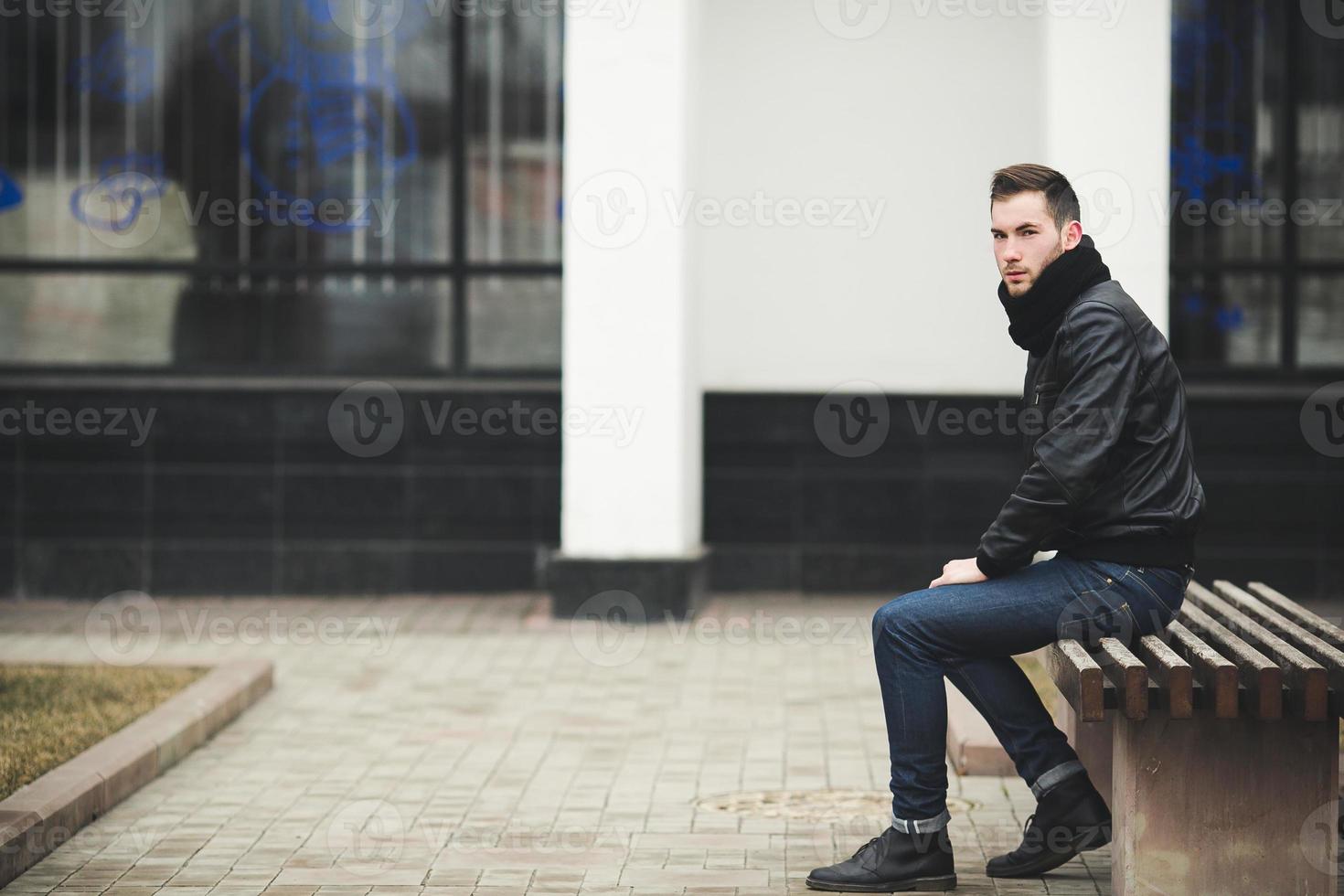 ein mann in jeans und schwarzer jacke sitzt auf einer bank foto