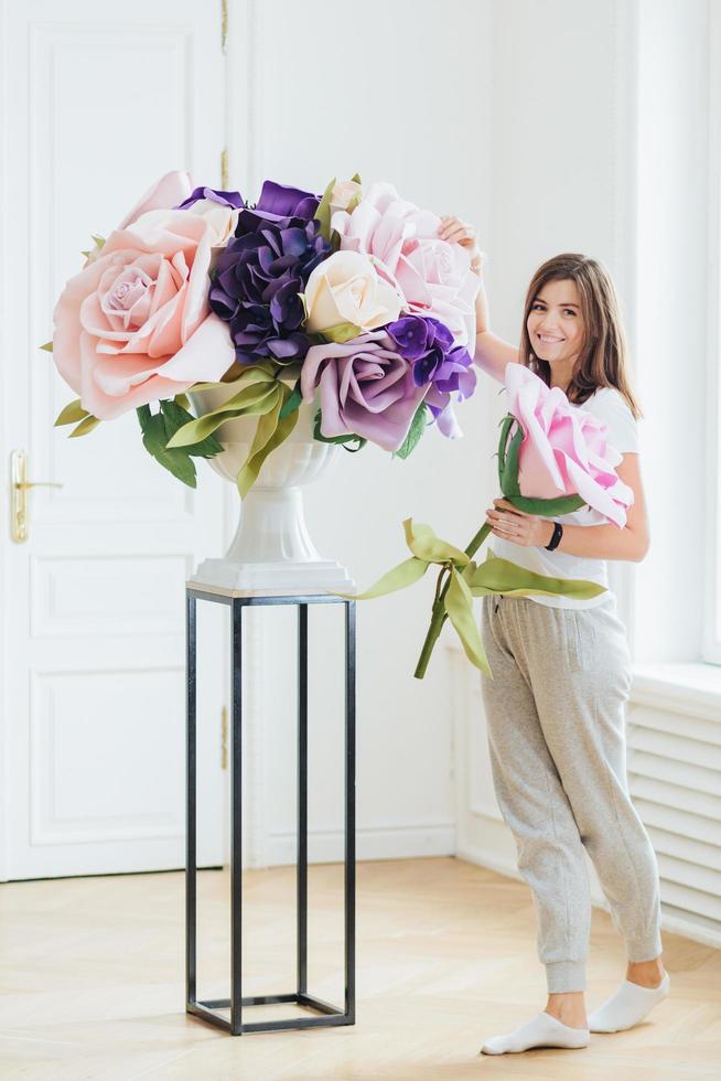 Ganzkörperfoto einer zufriedenen brünetten europäischen Frau in Freizeitkleidung, macht einen Blumenstrauß, posiert in einem geräumigen Raum. junge Geschäftsfrau besitzt Blumenladen. Innenaufnahme foto