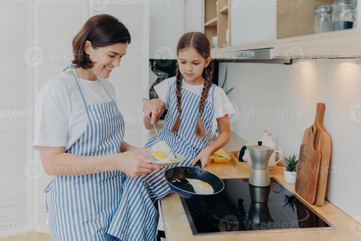 Foto von fröhlicher, beschäftigter Mutter und ihrer Tochter, die neben dem Herd posieren, Frühstück für die Familie servieren, Eier in der Pfanne braten, Schürzen tragen, in der heimischen Küche posieren. mutter bringt dem kind bei, wie man leicht mahlzeiten zubereitet