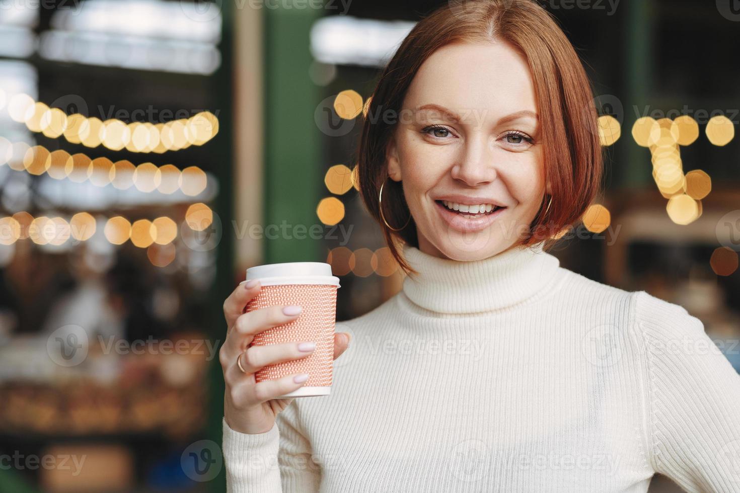 optimistische schöne frau mit gefärbten haaren, zufriedenem ausdruck, trägt einen rollkragenpullover, hält kaffee zum mitnehmen, posiert im außenrestaurant, hat ein schönes getränk, ist in bester stimmung, mag cappuccino foto