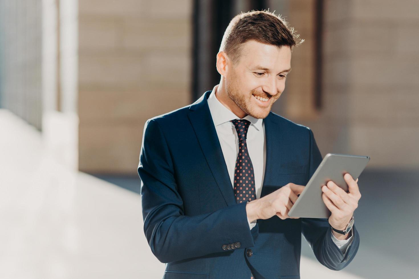 erfolgreicher männlicher unternehmer im formellen anzug, hält ein modernes tablet in der hand, prüft die dokumentation online, liest die notwendigen informationen für die erstellung des geschäftsberichts, nutzt die kostenlose wifi-verbindung. Menschen und Technik foto