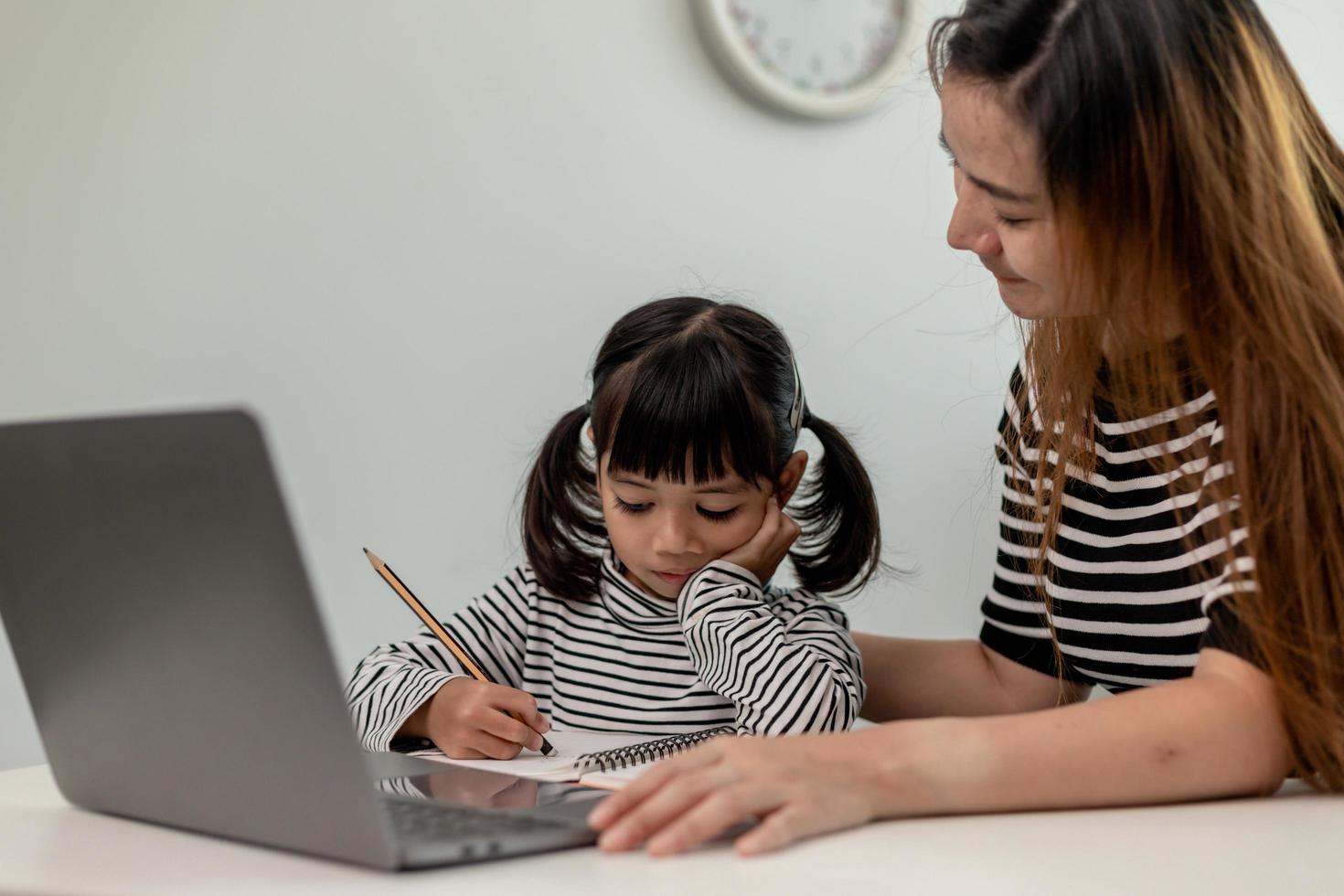 asiatisches kleines junges mädchen, das zu hause mit der mutter online-unterricht lernt. vorschulkind benutzt laptop-computer, macht hausaufgaben, homeschool vom schullehrer über digitales remote-internet mit unterstützung von mama. foto