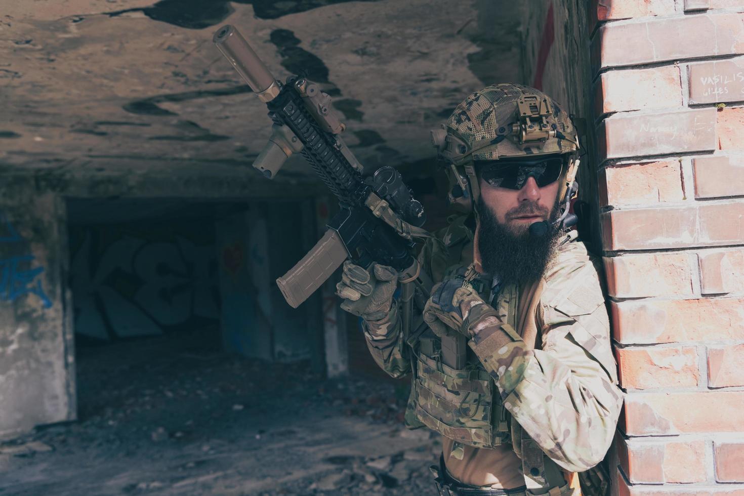 ein bärtiger soldat in uniform einer spezialeinheit in einer gefährlichen militärmission, archiviert seine waffe, während er sich vor der wand versteckt. selektiver Fokus foto