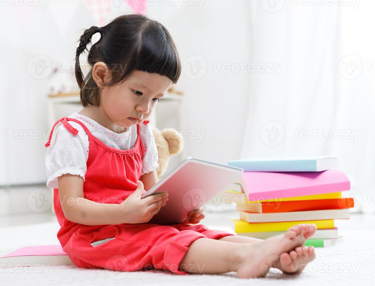 Vorschulmädchen, das Tablette schiebt. süßes kind, das mit teddybär liest. kleines Mädchen, das sich zu Hause, im Kindergarten oder im Kindergarten amüsiert foto