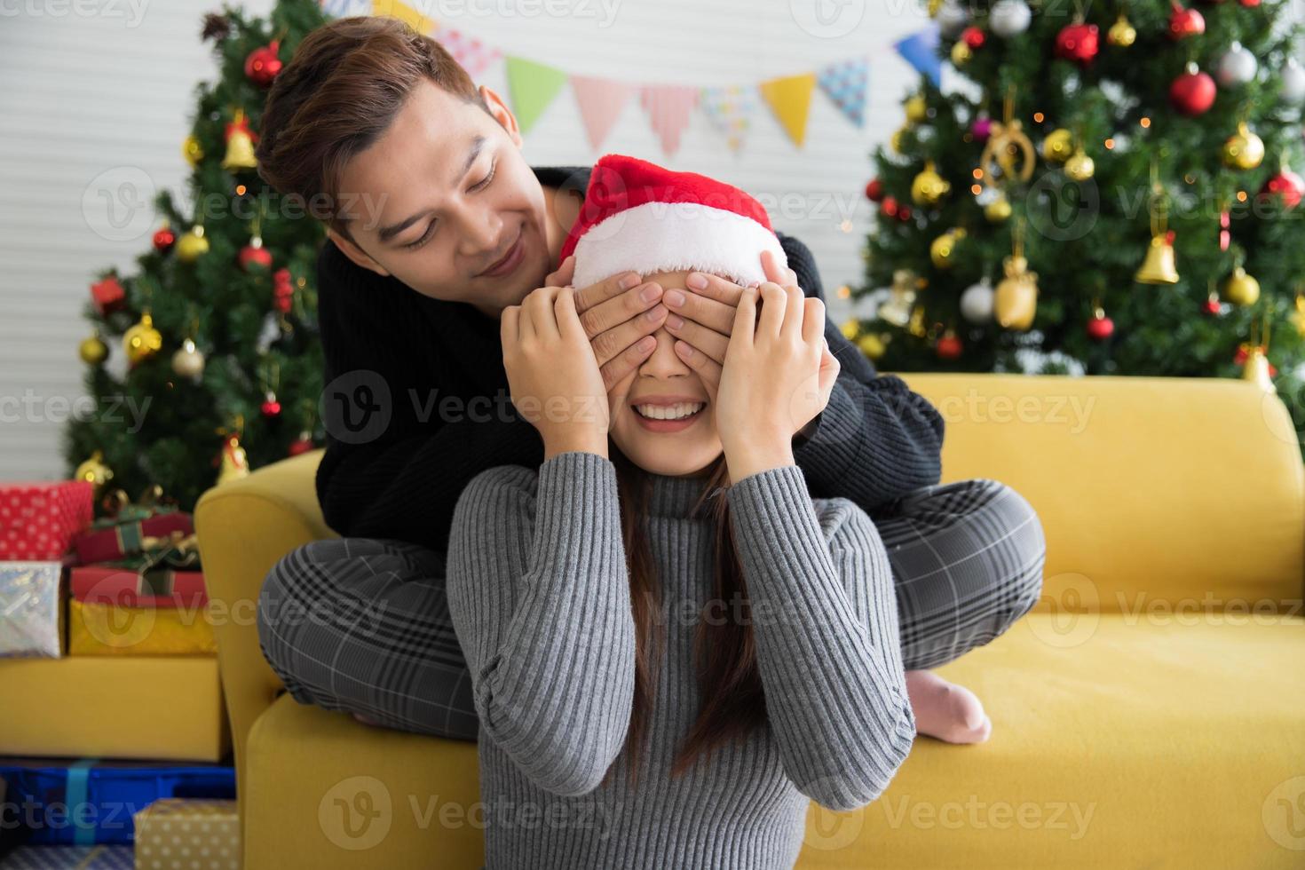 Schöner Mann, der eine Weihnachtsgeschenküberraschung für eine schöne Frau in Weihnachtsmütze mit geschlossenen Augen vorbereitet, Geschenkkartenkonzept foto
