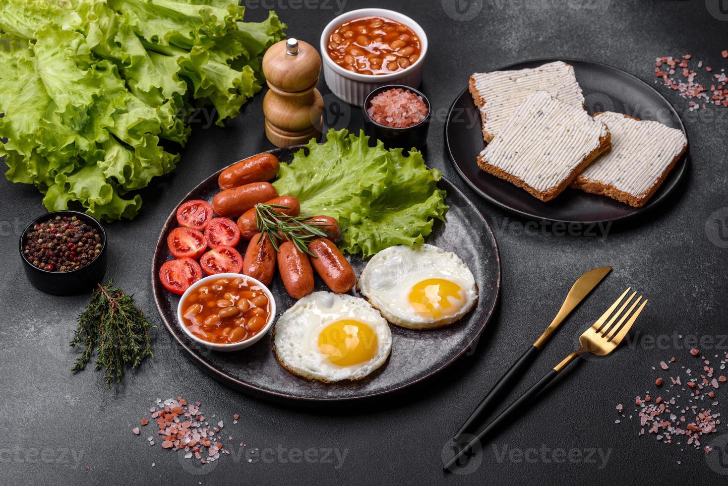 Traditionelles englisches Frühstück mit Eiern, Toast, Würstchen, Bohnen, Gewürzen und Kräutern auf einer grauen Keramikplatte foto