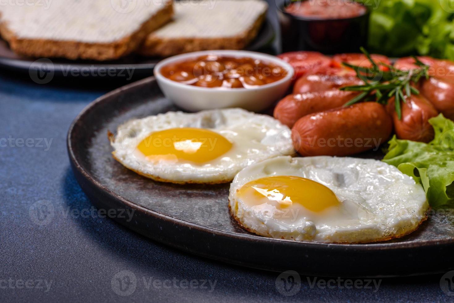 Traditionelles englisches Frühstück mit Eiern, Toast, Würstchen, Bohnen, Gewürzen und Kräutern auf einer grauen Keramikplatte foto