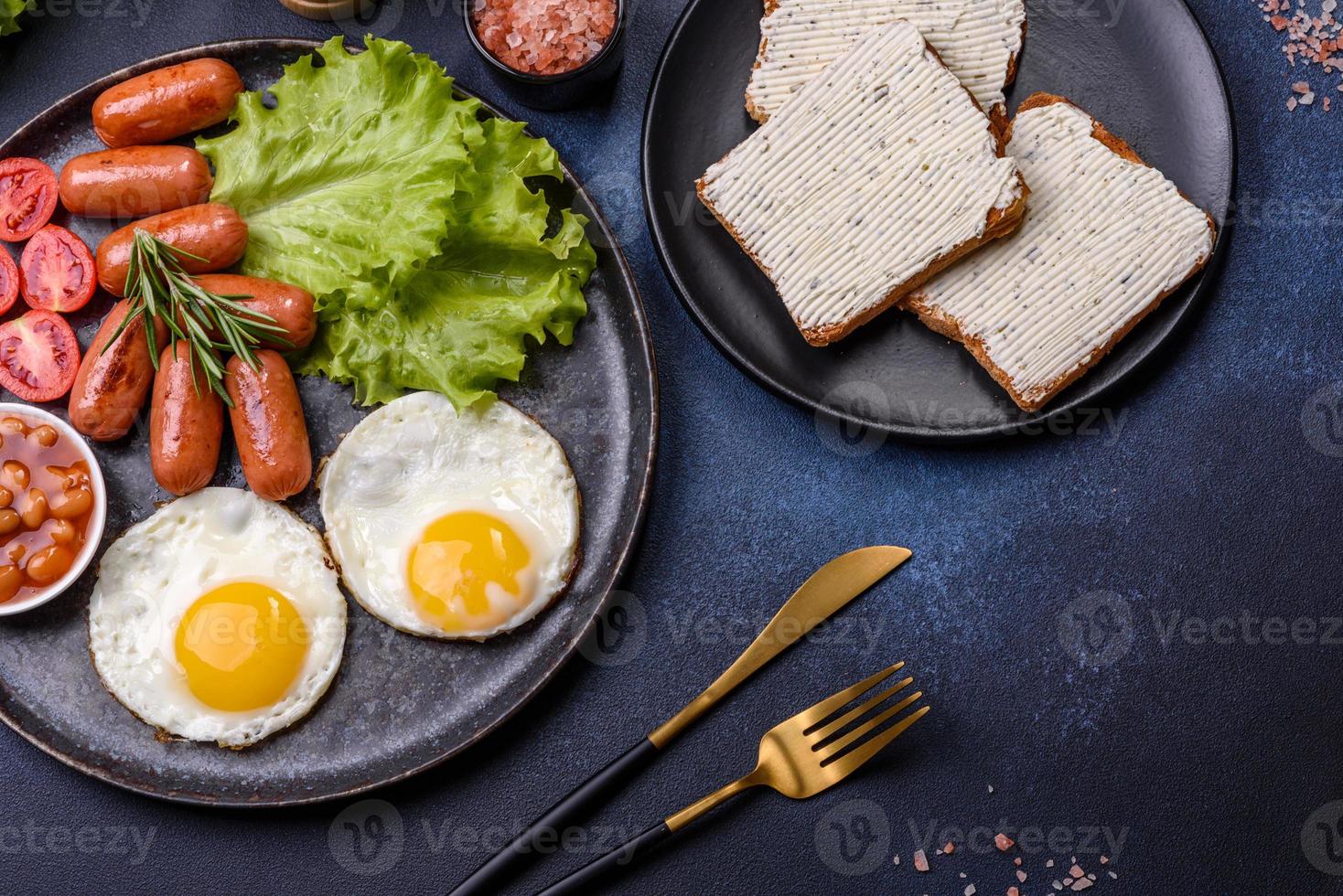 Traditionelles englisches Frühstück mit Eiern, Toast, Würstchen, Bohnen, Gewürzen und Kräutern auf einer grauen Keramikplatte foto