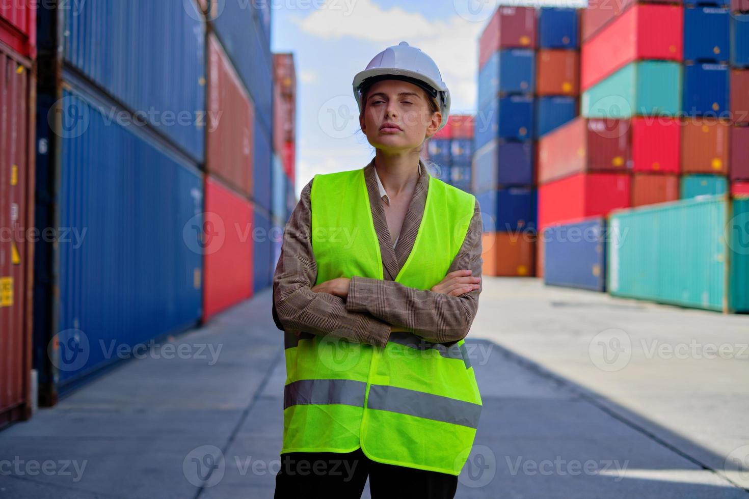 porträt einer professionellen weißen arbeiterin in sicherheitsuniform und mit verschränkten helmen und blick auf kameraarbeiten bei logistikcontainern, import- und exportfrachtschifffahrt, transportindustrie. foto