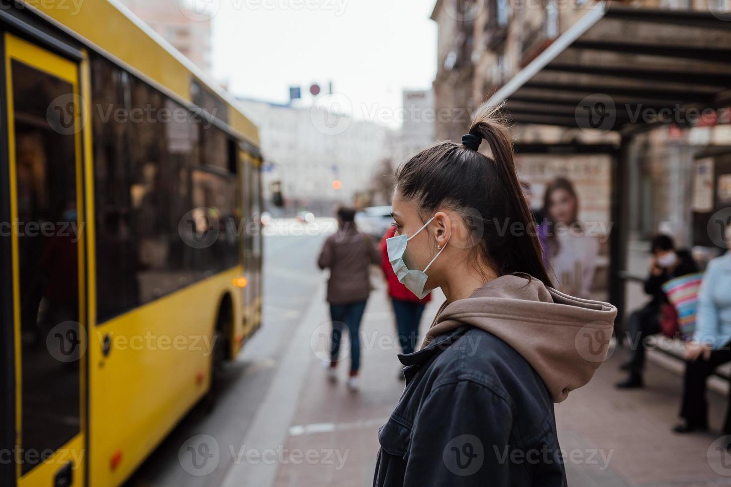 junge Frau mit chirurgischer Maske im Freien an der Bushaltestelle auf der Straße foto