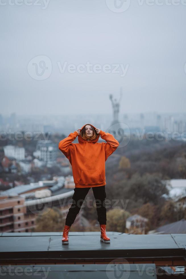 Mädchen in einer orangefarbenen Jacke posiert auf dem Dach eines Gebäudes im Stadtzentrum foto