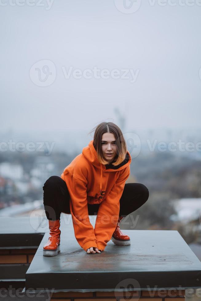 Mädchen in einer orangefarbenen Jacke posiert auf dem Dach eines Gebäudes im Stadtzentrum foto