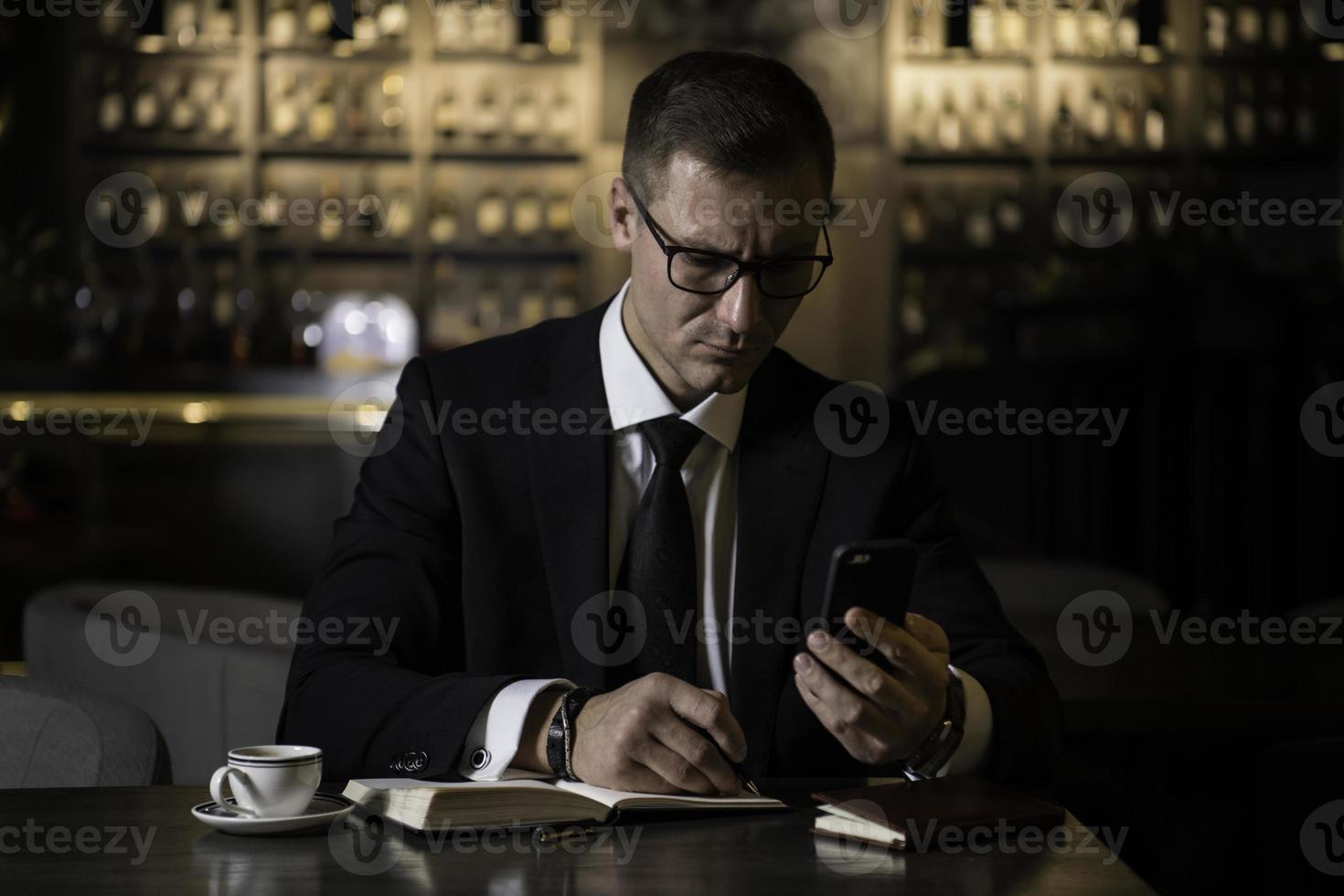 Porträt des ernsthaften gutaussehenden eleganten kaukasischen Geschäftsmannes, der am modernen Restaurant sitzt, sein Smartphone prüft und die Notizen in sein Notizbuch schreibt foto