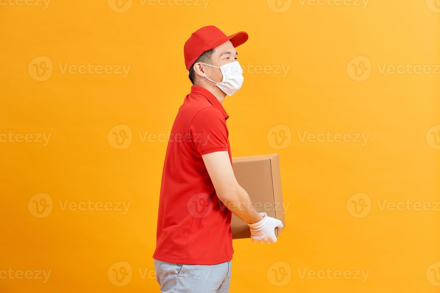 lieferbote mitarbeiter in roter kappe leeres t-shirt einheitliche gesichtsmaske handschuhe halten pappkarton isoliert auf gelbem hintergrund foto