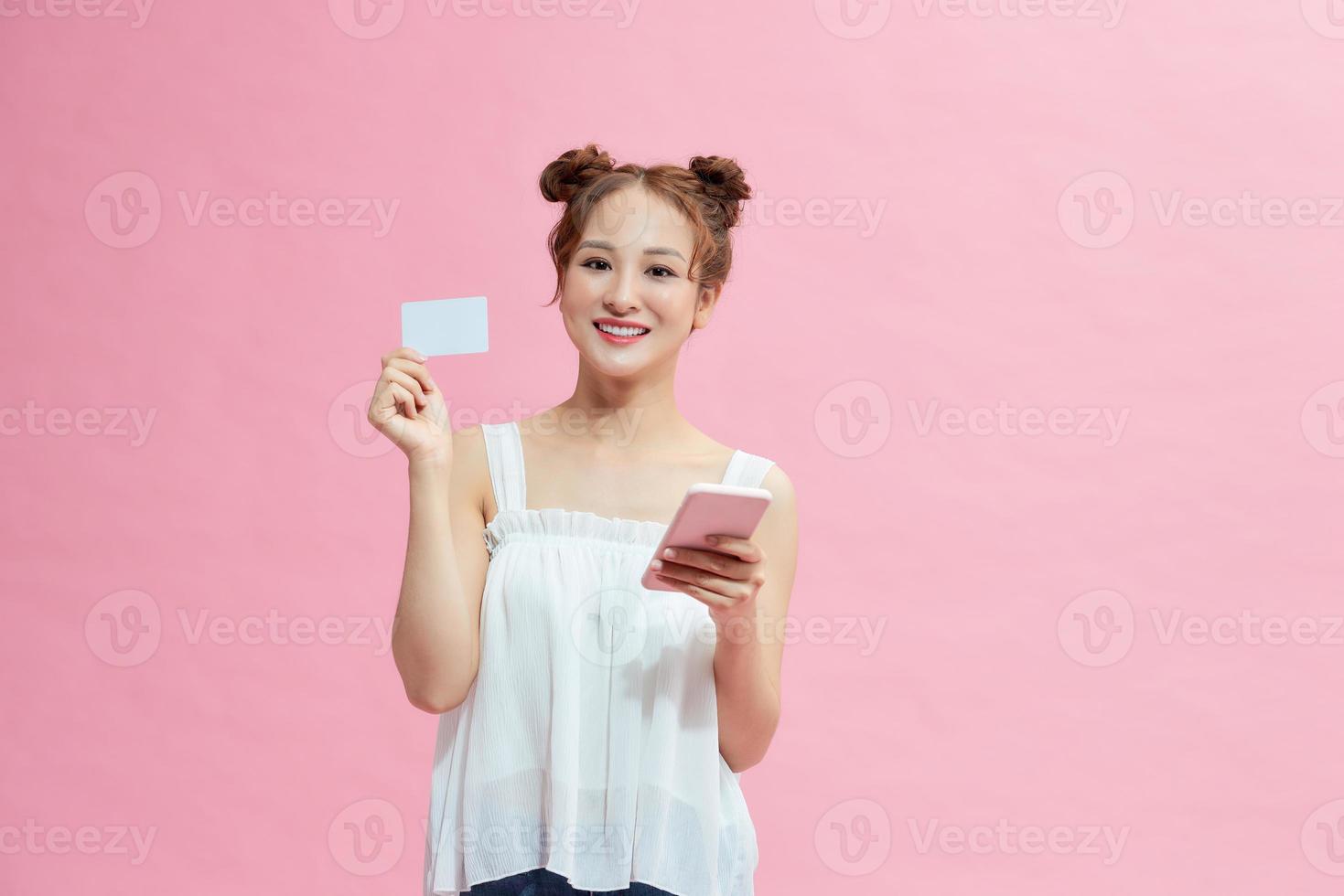 Foto einer erfreuten jungen Frau, die isoliert über einem rosafarbenen Wandhintergrund mit einem Mobiltelefon posiert, das eine Debitkarte hält.