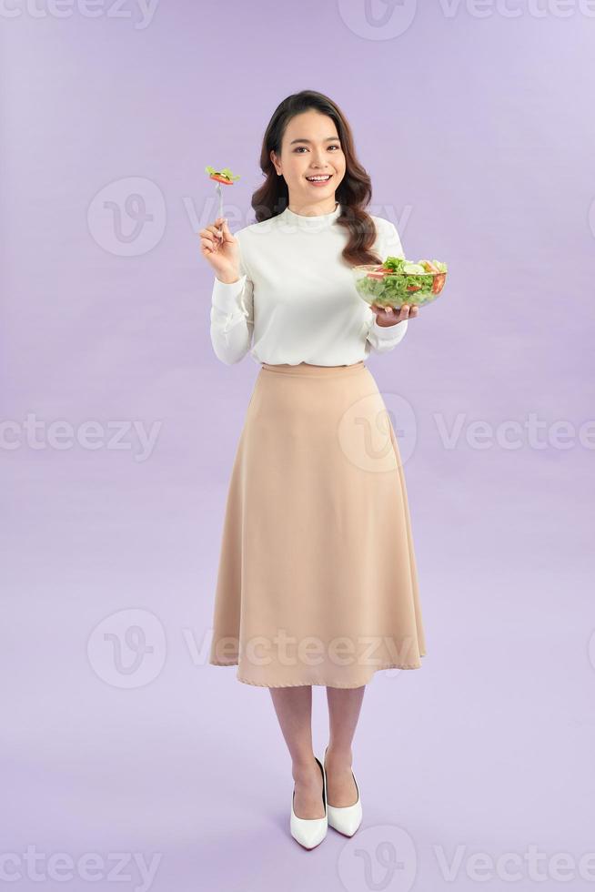 Porträt eines fröhlichen, verspielten Mädchens, das frischen Salat aus einer Schüssel auf violettem Hintergrund isst foto