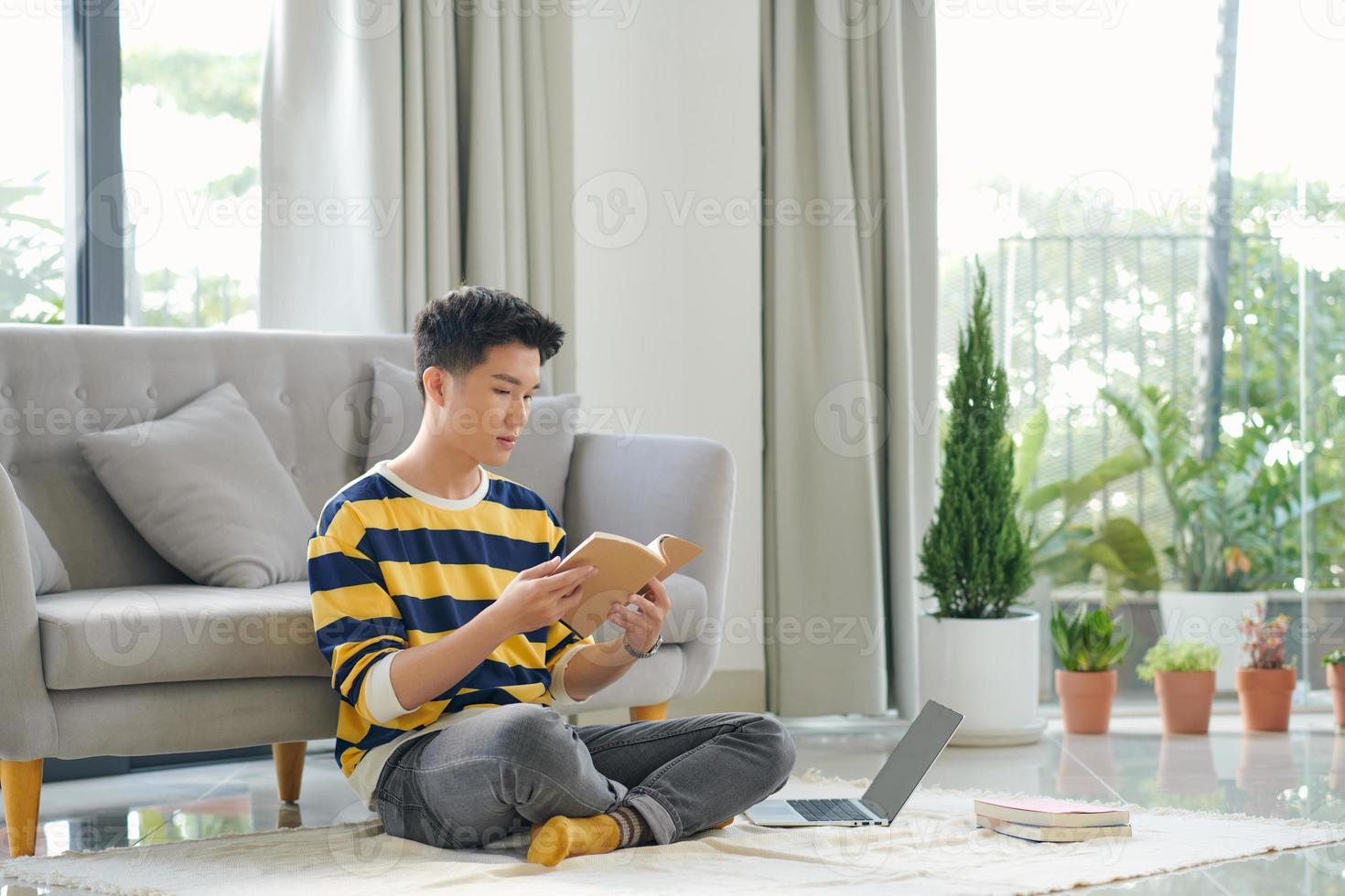 asiatischer mann sitzt mit einem buch und einem laptop auf dem boden foto