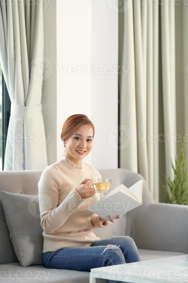 hübsche junge frau, die sich entspannt, ein buch auf einem bequemen sofa zu hause lesend foto