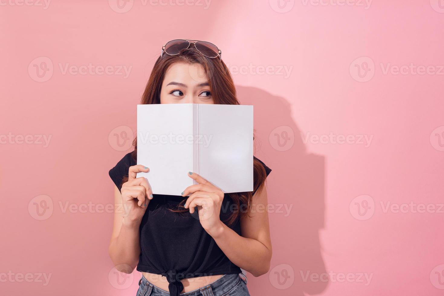 Porträt eines hübschen jungen Mädchens, das sich hinter einem offenen Buch versteckt und isoliert über rosafarbenem Wandhintergrund wegschaut foto