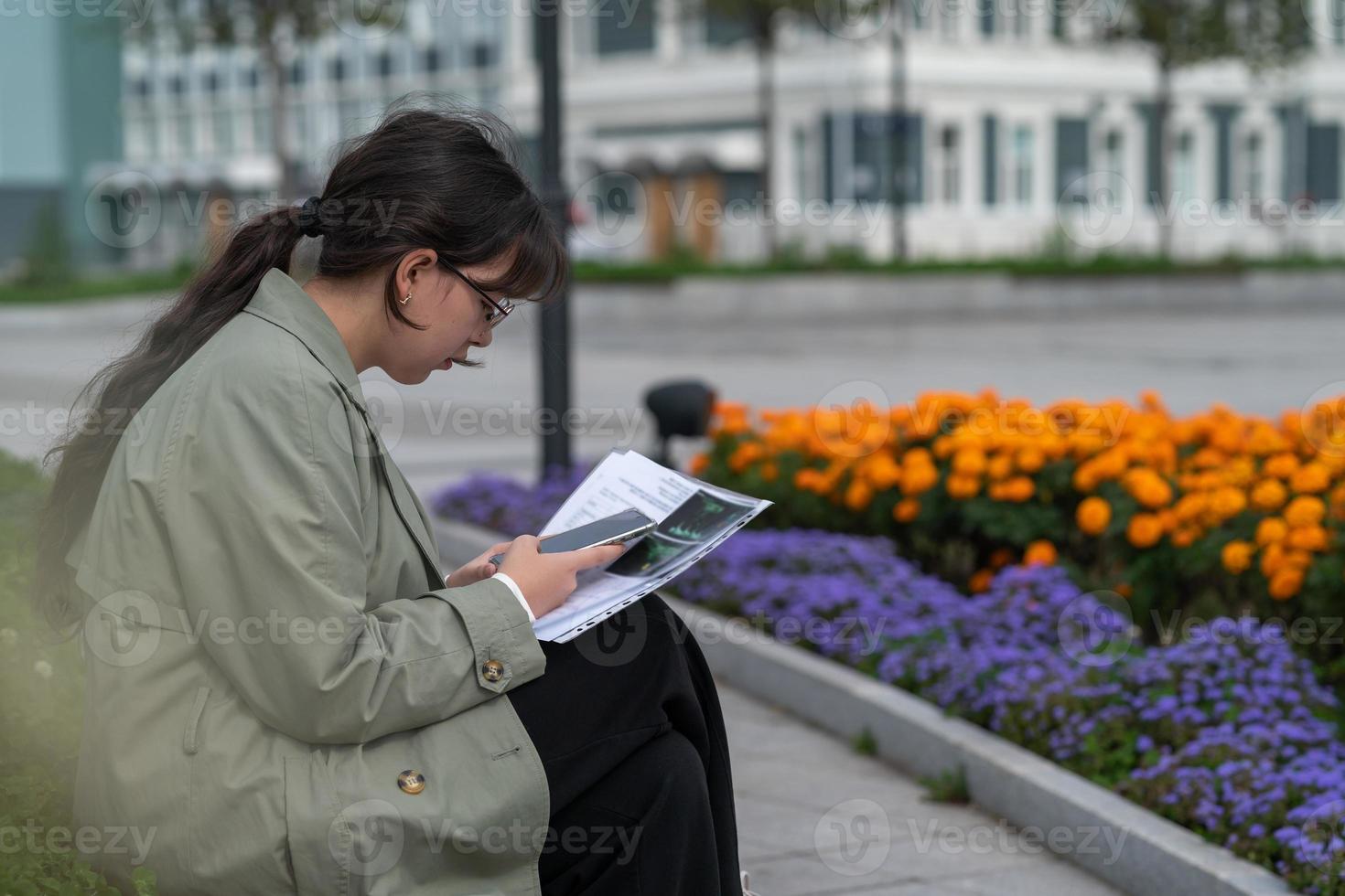 ein mädchen sitzt auf einer bank auf der straße mit dokumenten in den händen und benutzt ihr telefon foto