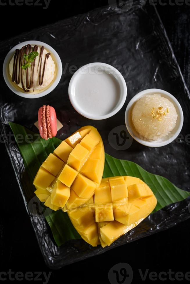 mango klebriger reis, kokosmilch, eis und macarons auf einem schwarzen teller beliebte thailändische desserts im sommer foto