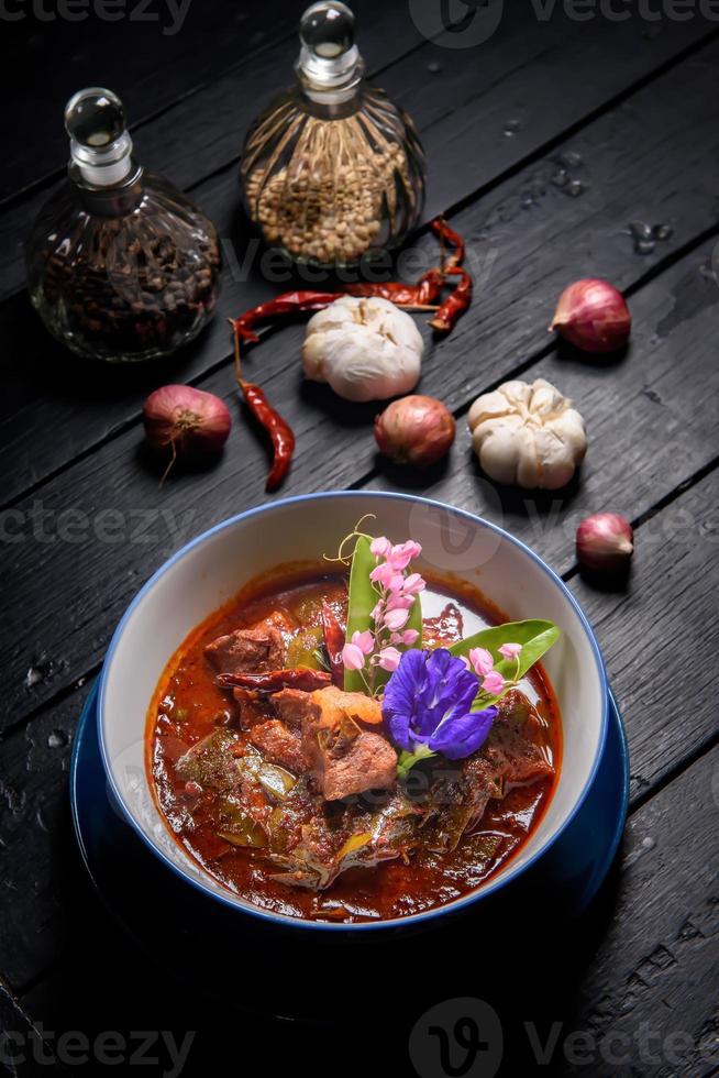 Rotes Curry aus Schweinefleisch, serviert und dekoriert mit Gemüse, Kräutern und Gewürzen auf rustikalem Hintergrund - thailändisches Essen foto