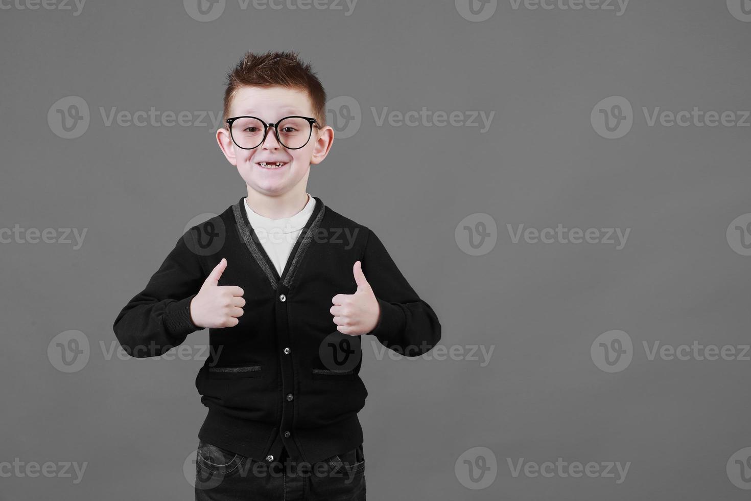 wie. Porträt eines glücklichen kleinen Schuljungen mit Brille, der in die Kamera lächelt und eine Daumen-hoch-Geste macht, die ein cooles Zustimmungszeichen zeigt. Indoor-Studioaufnahme isoliert auf grauem Hintergrund foto