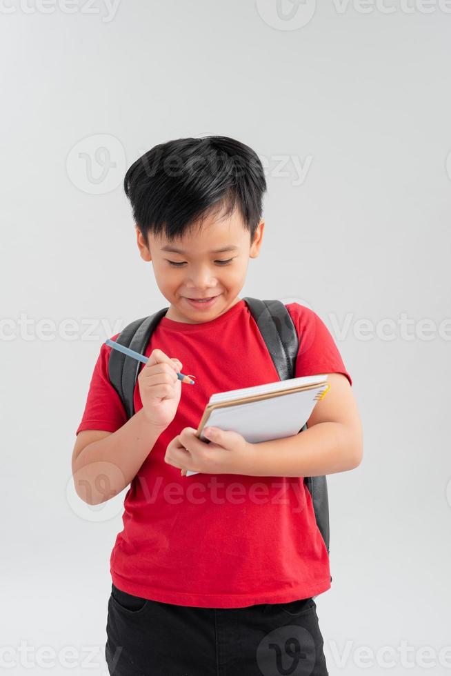 Schuljunge mit Rucksack, der etwas auf sein Notizbuch schreibt, isoliert auf weiß. foto