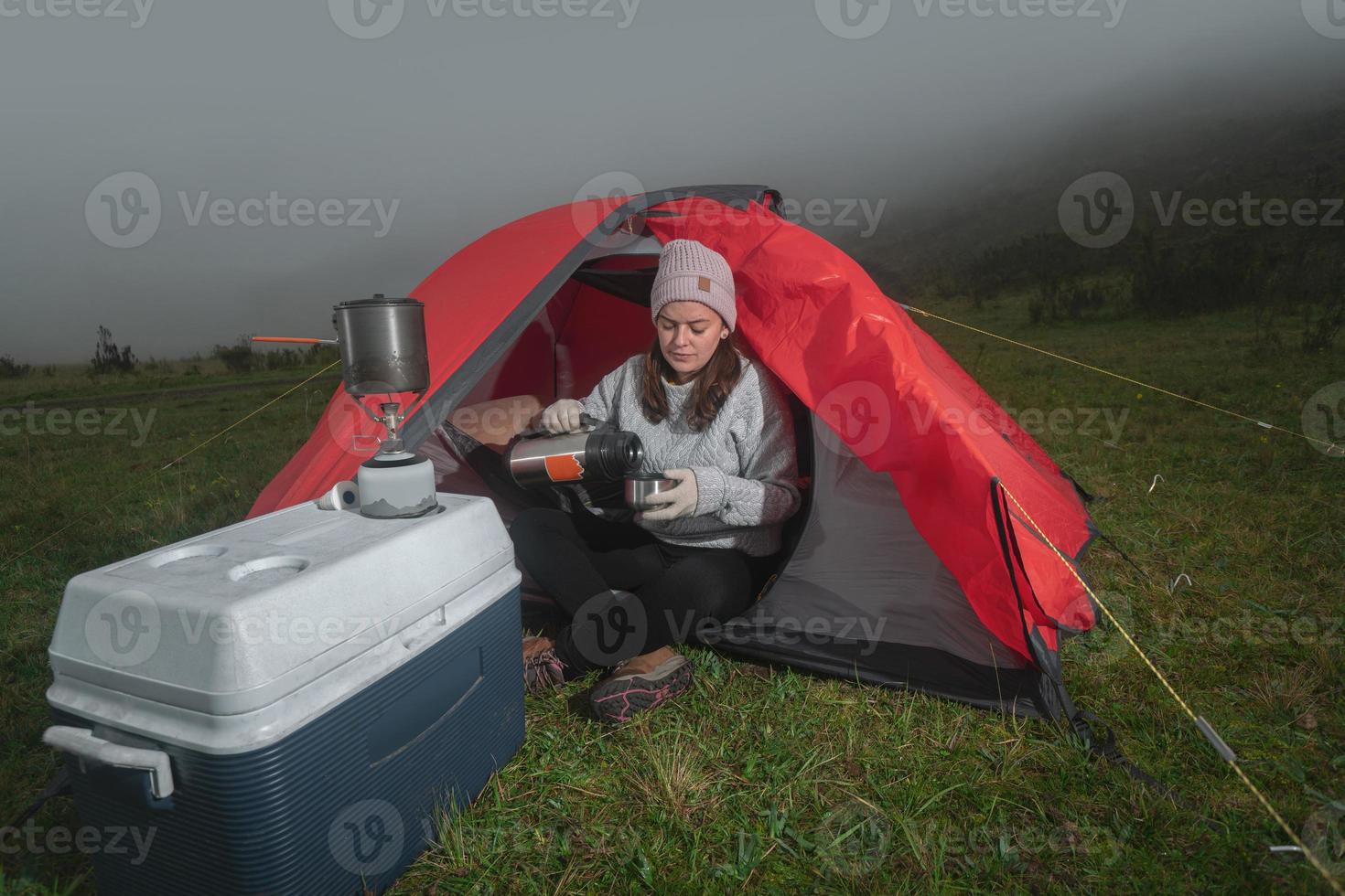 Schöne lateinische Frau, die vor ihrem Zelt sitzt und Tee aus ihrer Thermoskanne bei einem bewölkten Sonnenaufgang trinkt foto
